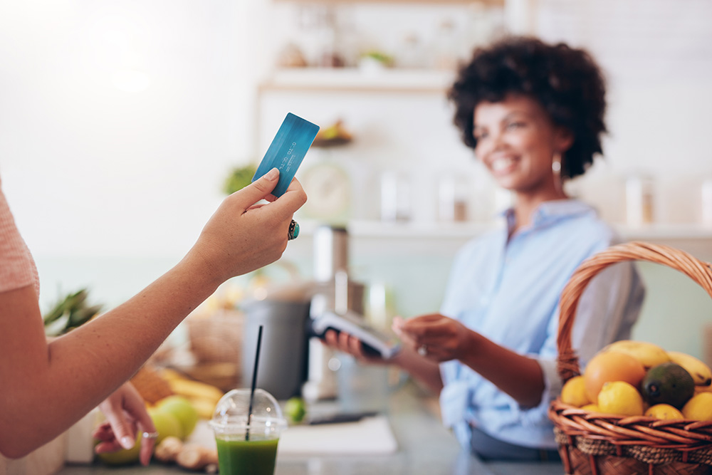 A blurred out photo of a smiling woman standing behind a produce counter. A hand holding a credit card is in the foreground. 