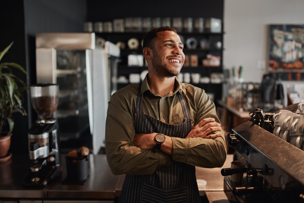 A man stands in a coffee shop, wearing an apron. He is smiling and staring at the corner. 