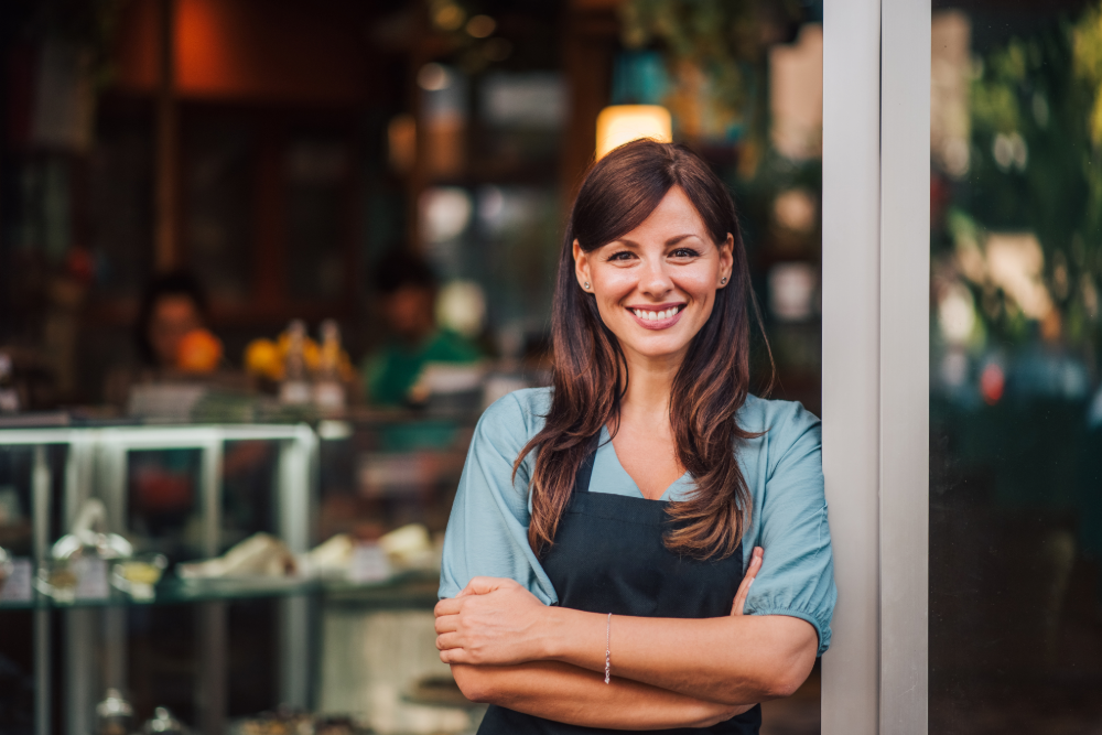 A woman in an apron stands in front of a window, smiling. 