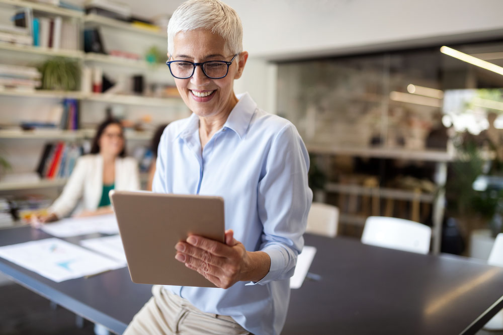 Elderly woman leans against a desk. She is wearing glasses and smiling at a tablet she is holding in her hand.