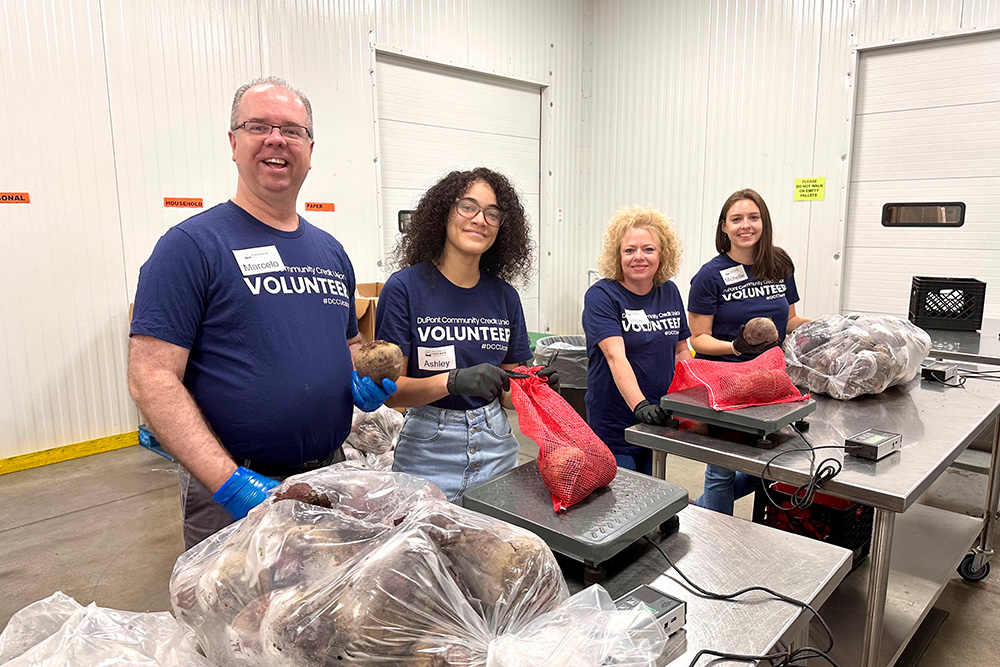 A group of DCCU employees pose behind a table at the Blue Ridge Area Food Bank. They are all wearing navy blue 'Volunteer' shirts. 