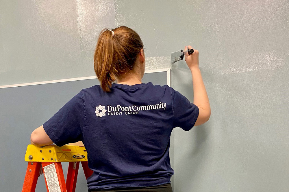 Female DCCU employee stands on a ladder with her back facing the camera, holding a paint brush and painting a wall.
