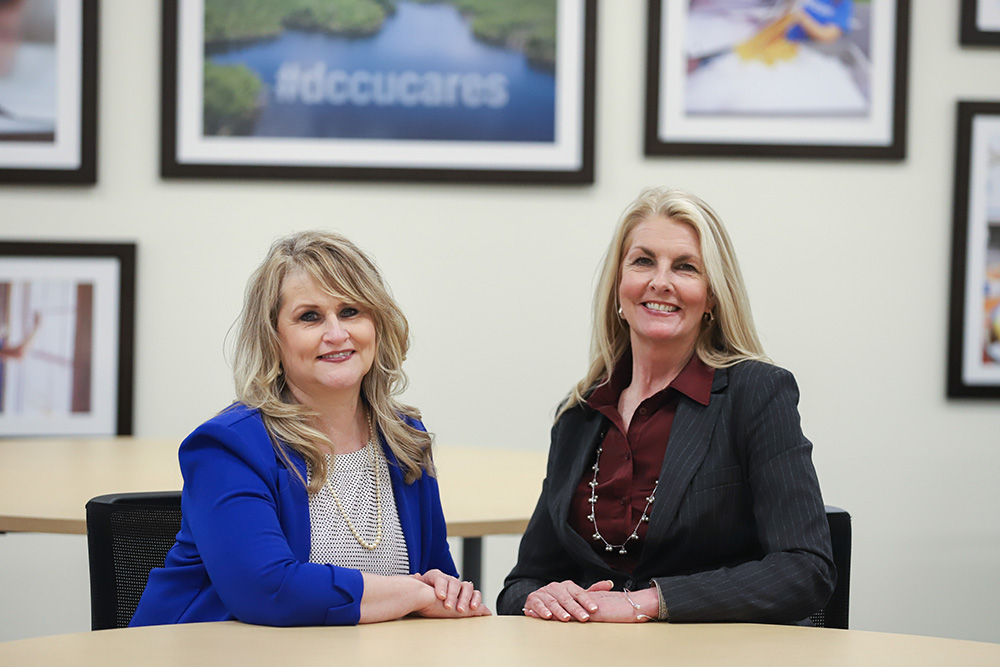 Sarah Landram and Margaret Hersh (l-r), of the DCCU Cares Foundation sit at a table, smiling for the camera. 