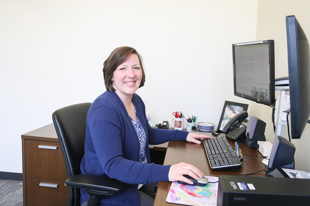 Woman at desk, smiling, with hand on mouse at computer