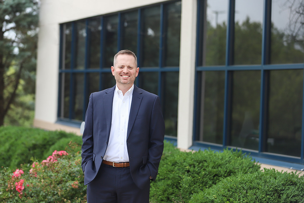DCCU's Senior Vice President, Chief Retail Officer, Jacob Larew, stands outside and smiles for the camera in front of a row of bushes. 