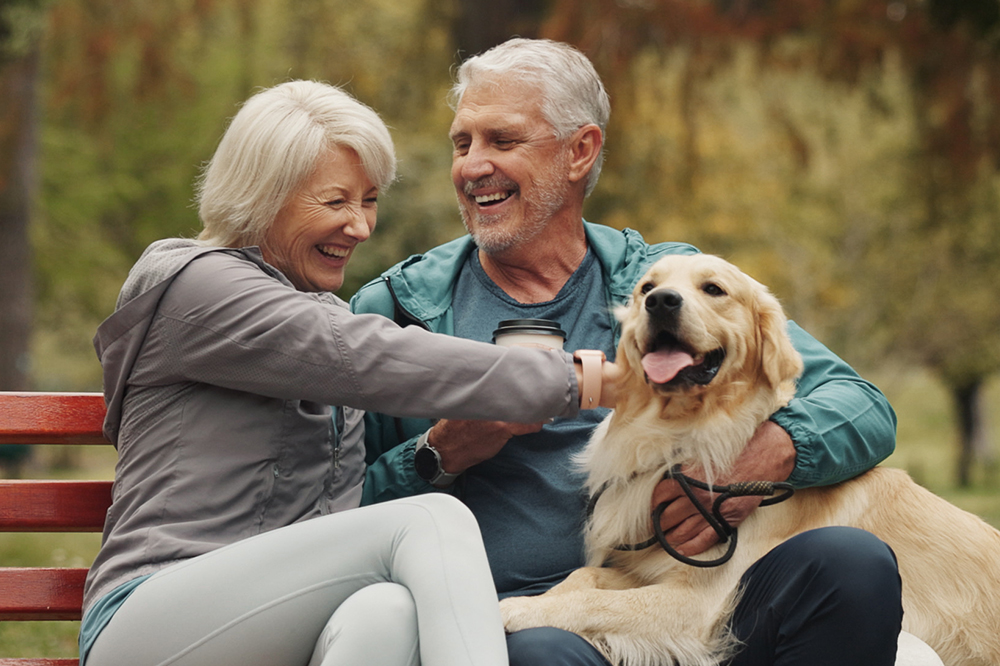 Man and woman sit on a bench outside, petting a dog