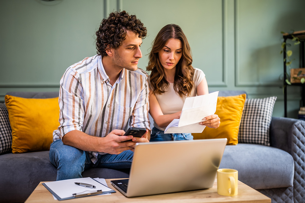 A young man and woman sit on a colorful couch while looking at a laptop. The man is holding a smartphone and the woman is holding a sheet of paper. It appears as though they are doing paperwork of some sort. 