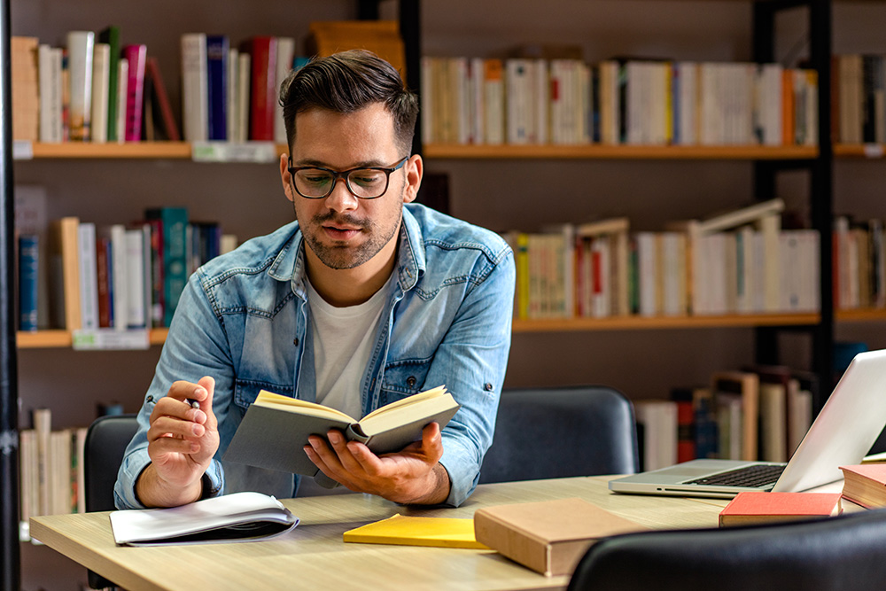 A young man sits at a table in the library, holding a book in one hand a pen in the other. 