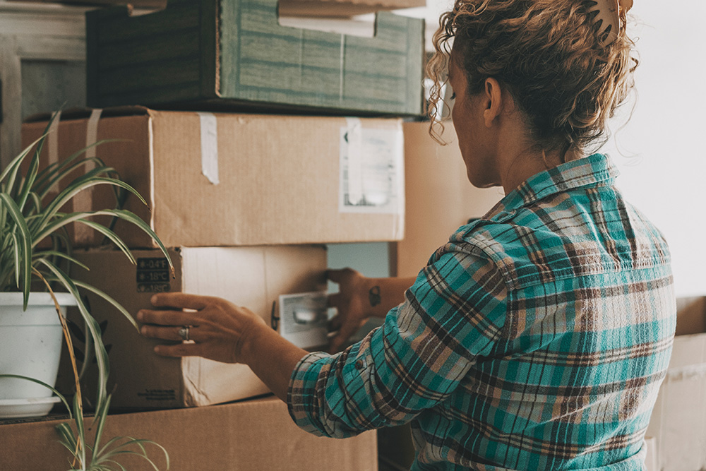 Image of the back of a woman touching a stack of cardboard boxes. She is wearing a blue plaid shirt, and has curly brown hair clipped high on her head. It appears as though she is moving. 