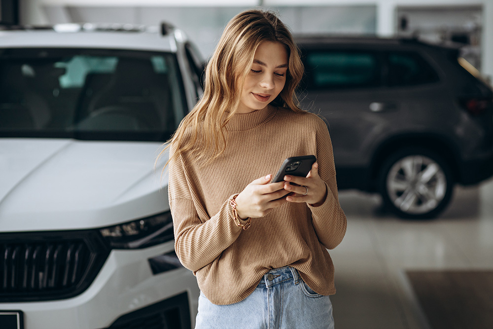 A young woman looks at a smartphone she is holding as she stands in the middle of a car dealership.
