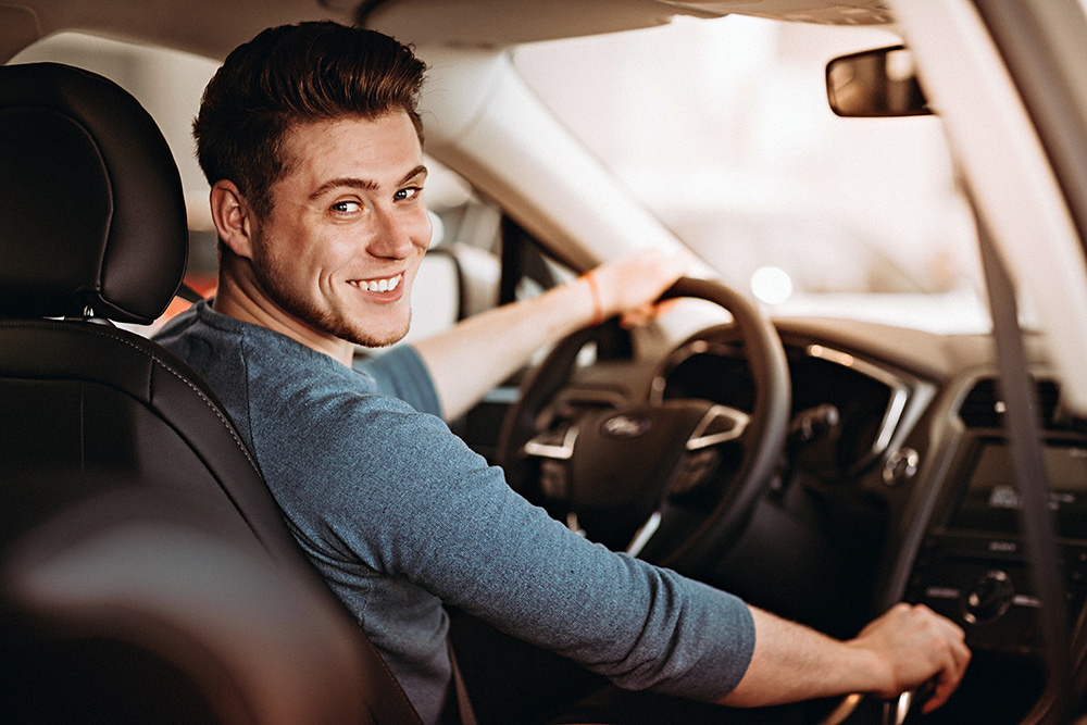 A young man sits in the driver seat of a vehicle. He is half turned around and smiling at the camera. 