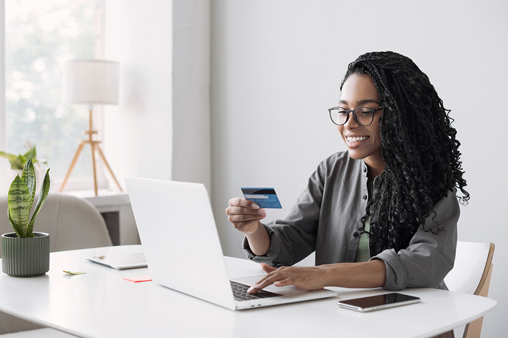 A young African American woman wearing glasses sits in front of  a laptop computer, holding a plastic credit card. She is smiling at the screen and using the computer keyboard. 