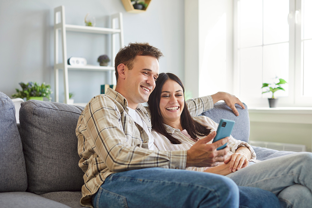 A young man and his wife sit on a sofa, looking at a smartphone he is holding. They are smiling. 