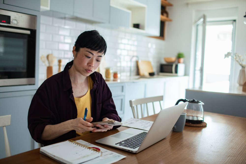 A woman sits at a kitchen table with a laptop, notebook, and papers in front of her. She is holding a pen and a calculator while looking down at the calculator. 