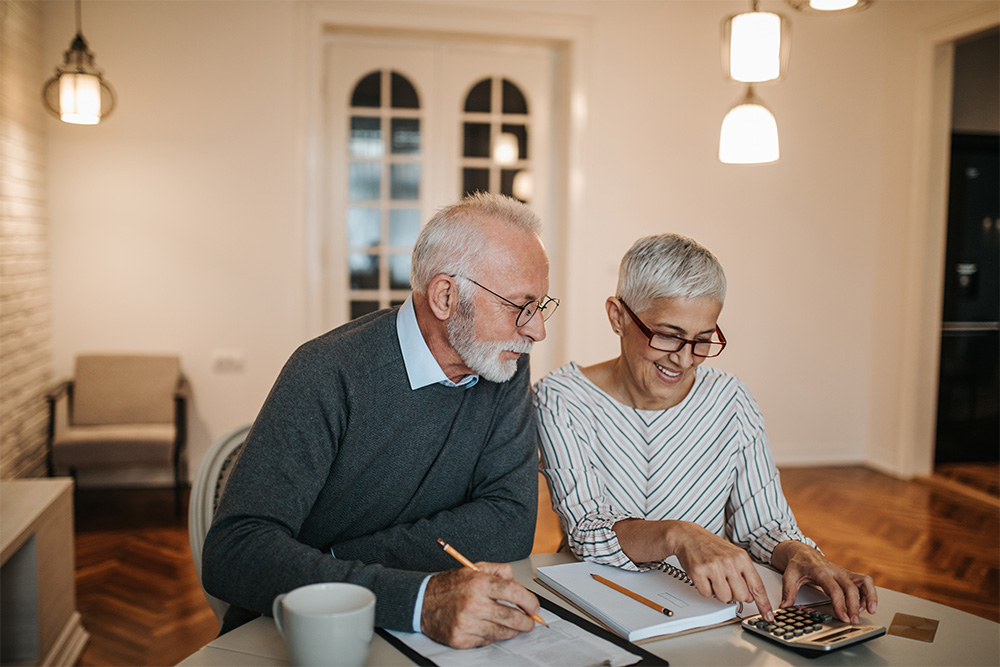 An elderly man and woman wearing glasses sit at a table, calculating and writing.