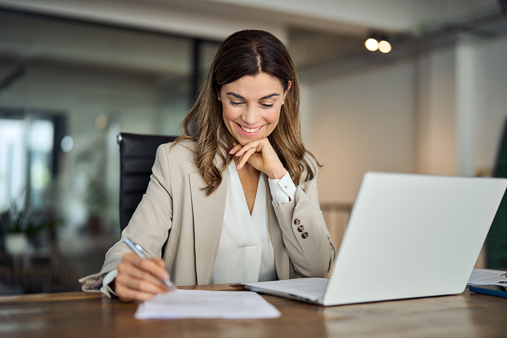 Woman in business suit sits at a table, writing on a piece of paper with a laptop in front of her. 