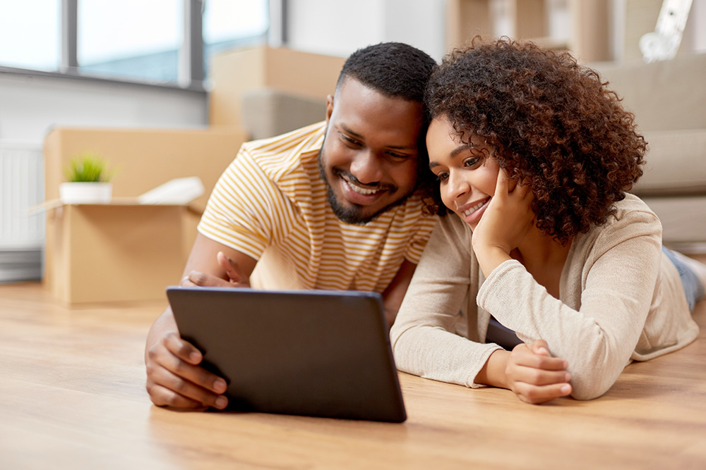 A man and his wife lay on their stomachs on a hardwood floor. They are smiling and looking at a tablet on the floor. 
