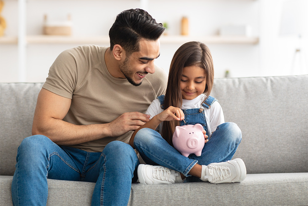 A man and and little girl sit on a couch. The little girl is holding a pink piggy bank and placing a coin inside it.