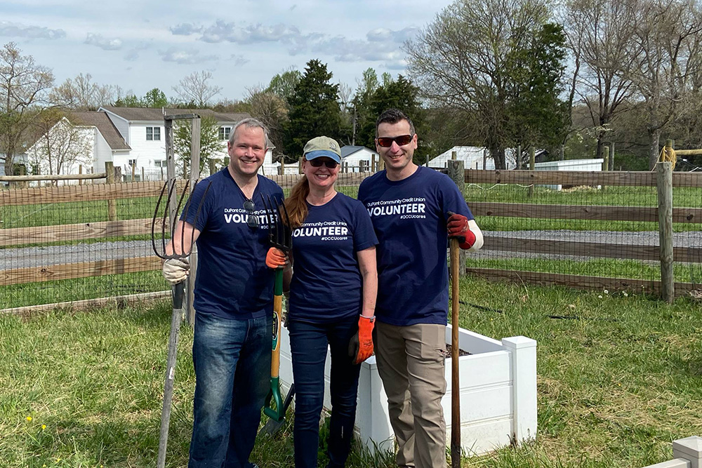 Three DCCU employees stand in a field, holding farming tools. They are volunteering. 
