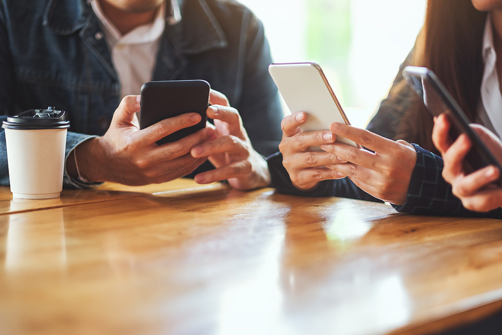 Three people sit at a table, with their phones out. They are all touching the phones. 