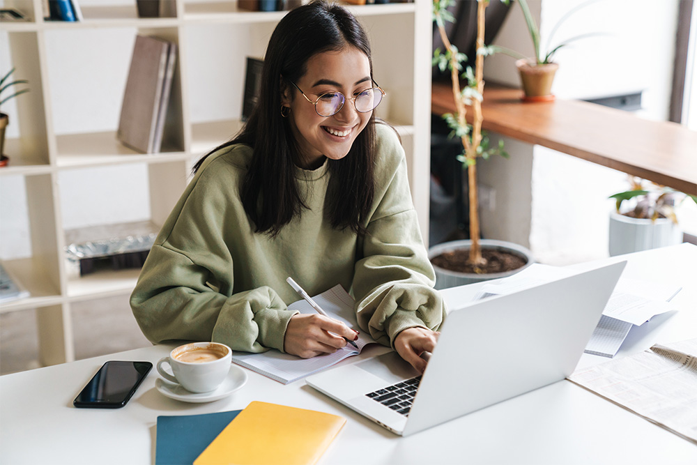 Image of smiling young woman holding a pen, using a computer, and writing things down in a notebook