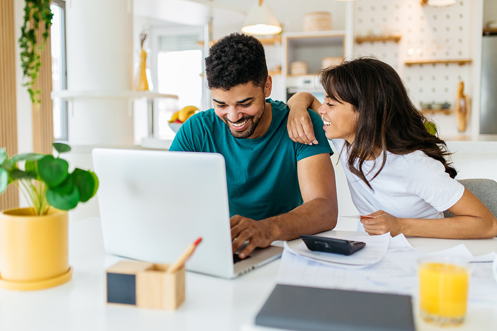 A young man and young woman sit behind a table in a kitchen, smiling. They are looking at a laptop computer on the table. 