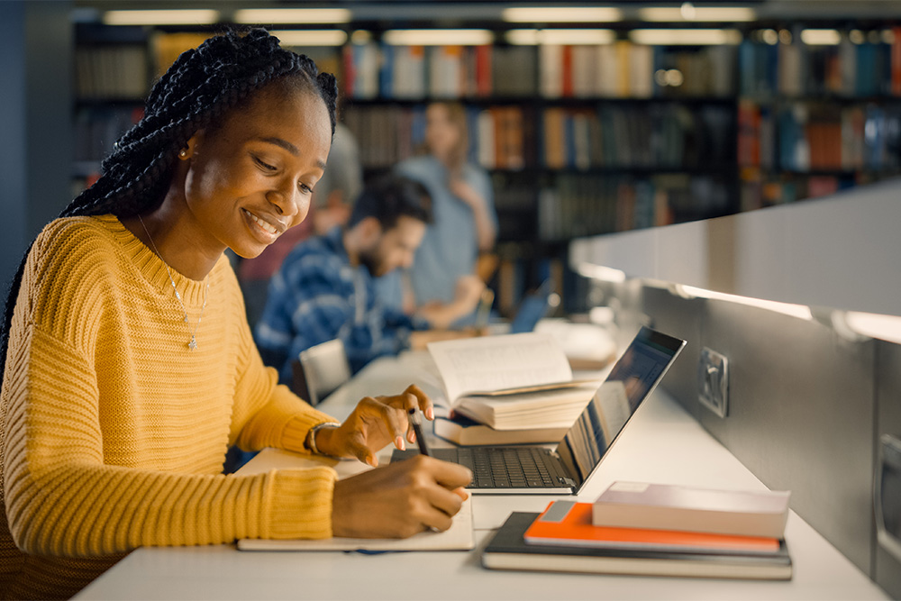 Young collegiate woman in library with laptop, writing in a notebook