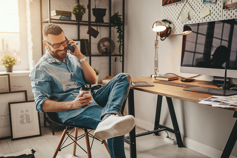 A man sits cross legged in an office chair with a cellphone to his ear. He is smiling and holding a to-go coffee cup
