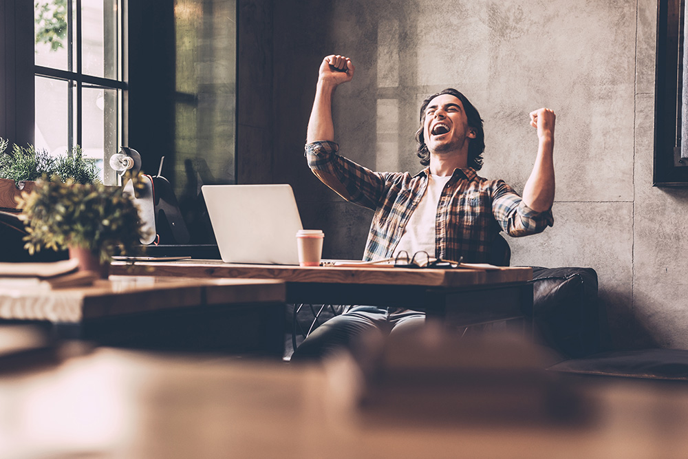 Man at desk excitedly raises arms in triumph.