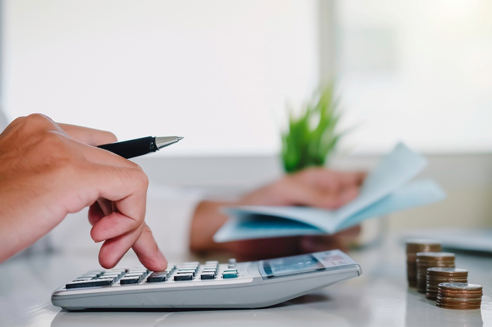 A close-up of a person's hand as they prepare their finances. They are holding a pen and typing on a calculator and in the other hand hold a sheet of paper. The background features stacks of coins and a table plant. 