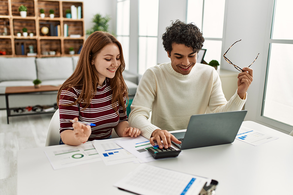 A young woman and young man sit at a table using a laptop and calculator. There are papers strewn about the table.