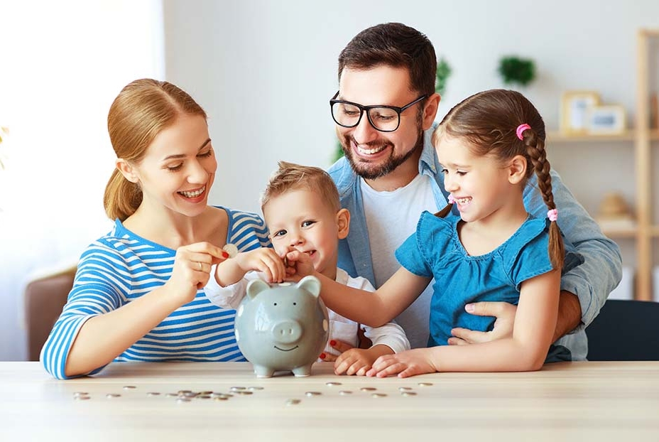 a woman, small boy, man, and young girl sit around a table, putting coins in a piggy bank