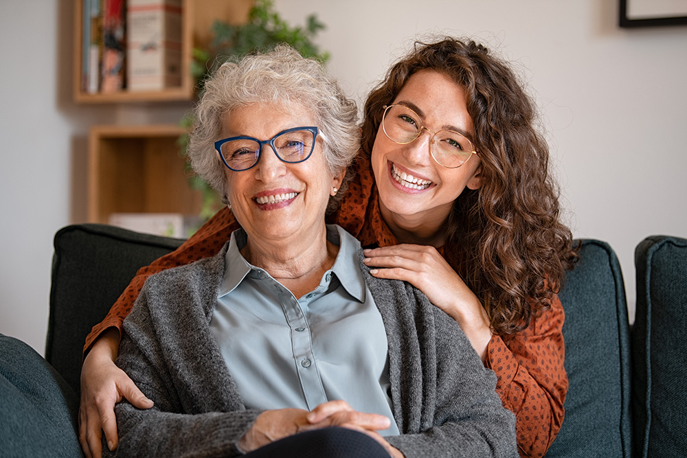 Young woman wearing glasses sits behind elderly woman wearing glasses with her arms draped over the elderly woman