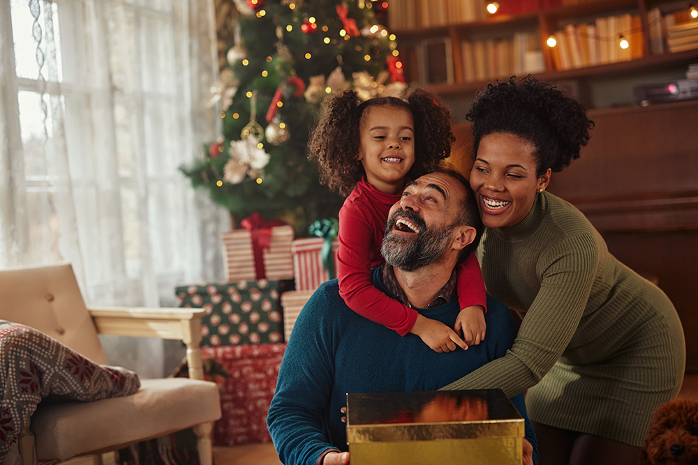A man holding a gold gift box is hugged from behind by a small little girl with pigtails and a woman with curly hair. 