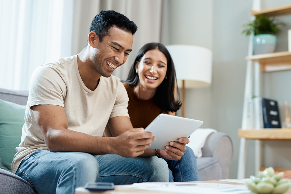 Man and woman sit on couch in living room, smiling. They are looking at an iPad or tablet together.