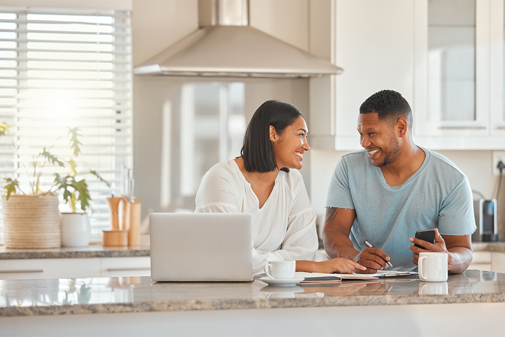 A man and woman sit at a kitchen counter, going over finances and sitting with a laptop. They are smiling at one another. 