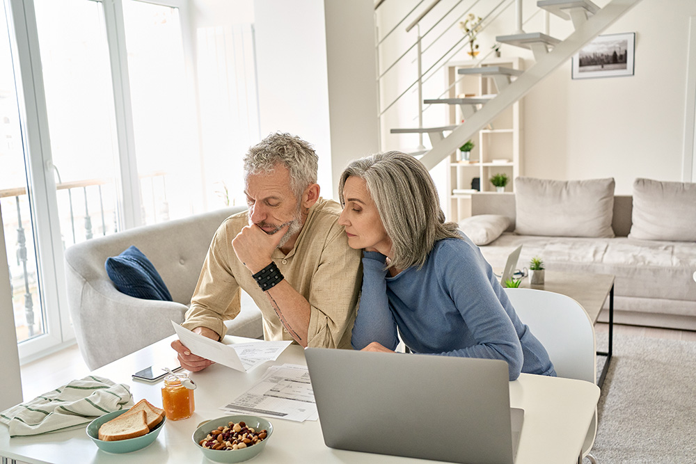 Elderly man and woman sit at a table in front of a laptop. They are studying a paper in the man's right hand.