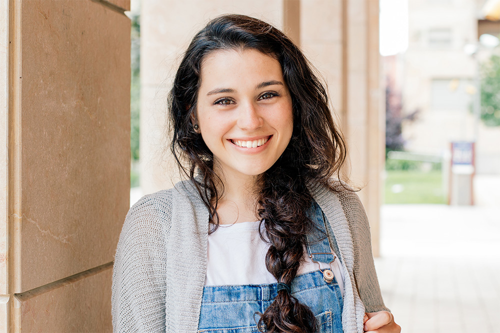 Young woman in college smiles at the camera, holding a backpack over her shoulder. 