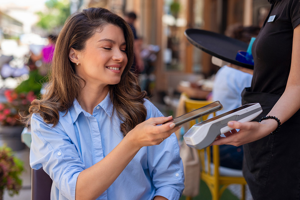 A woman with long wavy brown hair sits outside and pays for her purchase by tapping her phone on the terminal