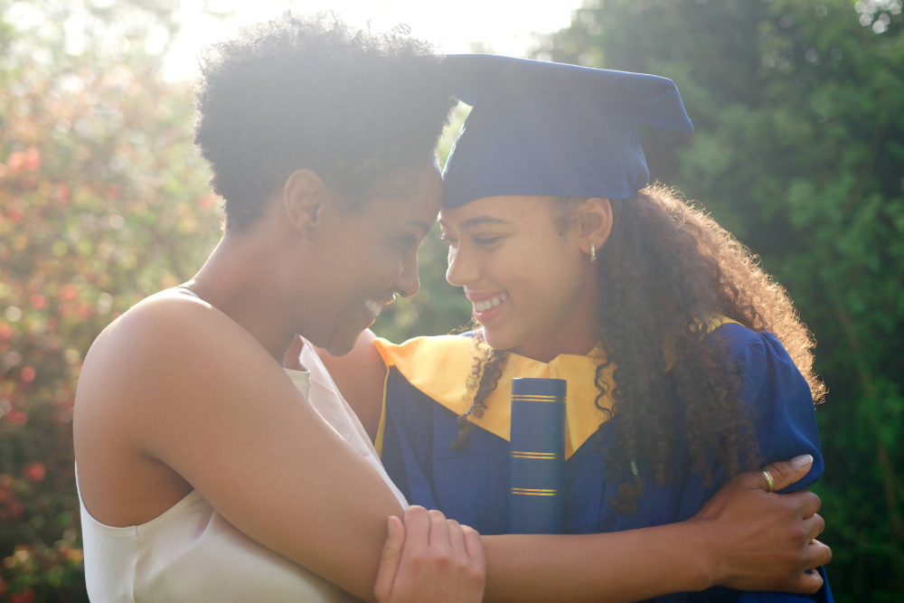 A young woman wearing graduation attire hugs an older woman. They are both smiling. 