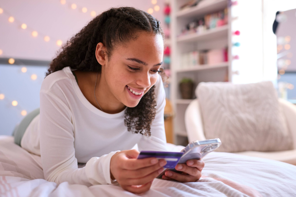 A young woman lays on her bed, holding a credit/debit card while smiling at her smartphone in her other hand. 
