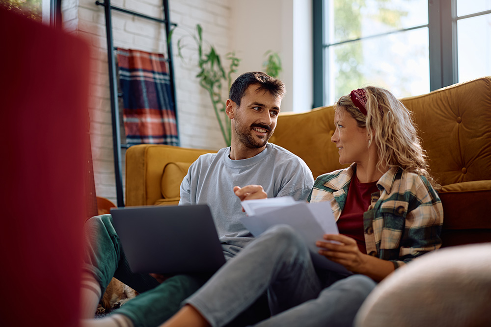 A man and woman sit on the floor in front of a yellow couch. The woman is holding a stack of papers while the man has a laptop on his lap. It appears as though they are talking about finances. 