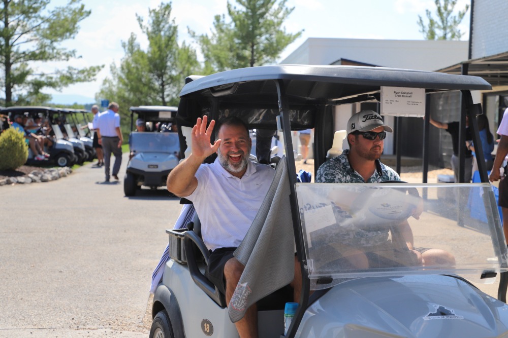 Two men sit in a golf cart. One of them is smiling and waving. 