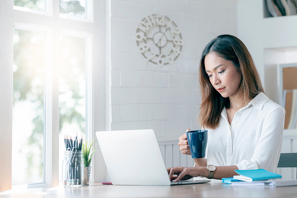 A woman sits at a laptop computer, holding a coffee cup in her hand. 