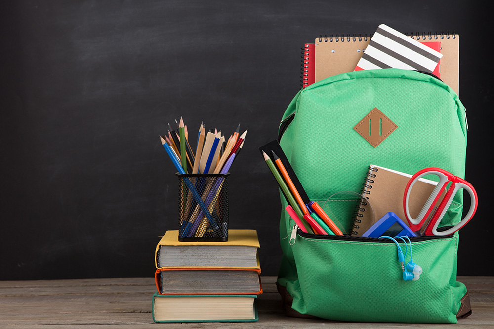 A green backpack full of school supplies sits next to a pile of books in front of a black chalkboard background. 