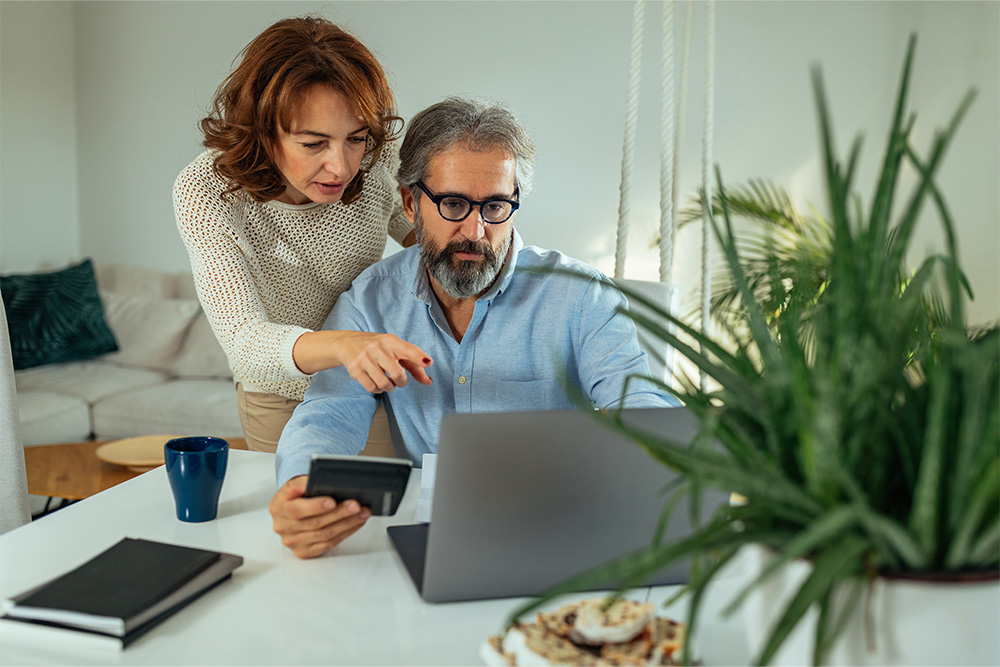 A woman and man sit behind a laptop computer. The woman is pointing to the screen as the man holds a calculator.