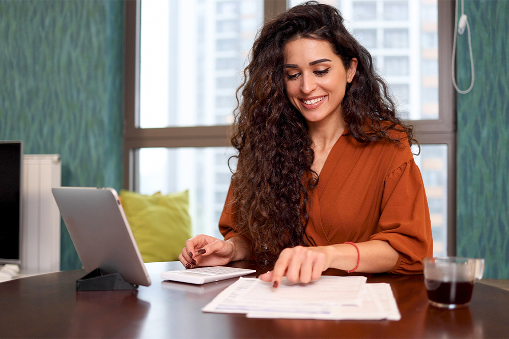 A woman with curly hair sits at a table looking over paperwork and smiling. 