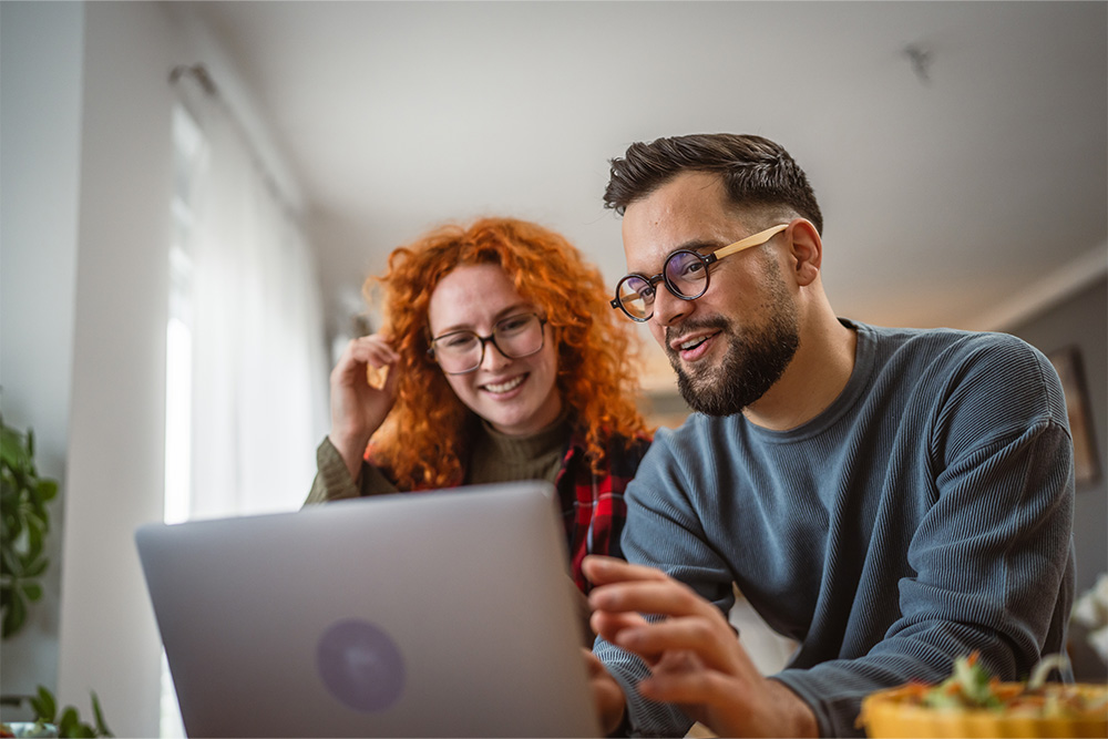 A woman and man sit behind a laptop, studying the screen. They are smiling. 