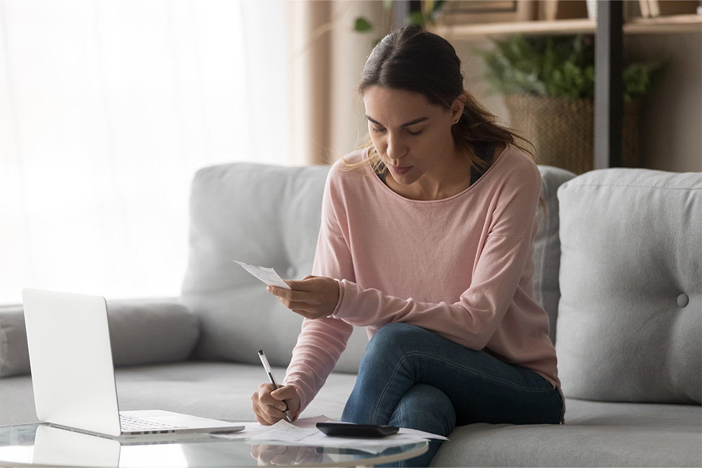 A woman sits on her couch with a pen in one hand and a receipt in the other. She is working on her finances. 