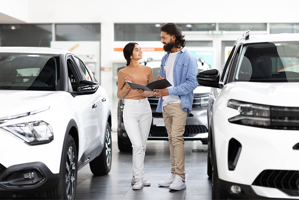 A young woman and man stand in a vehicle dealership showroom. The man is showing the woman paperwork in his hands. 